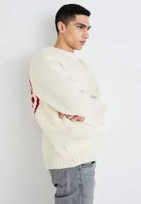 Pull en tricot crème avec un ourlet côtelé, présentant un motif géométrique rouge prononcé sur le côté gauche. Assorti à un pantalon gris.