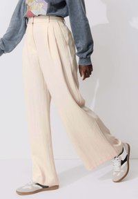 Pantalons plissés à jambes larges, de couleur rose pâle, fabriqués en tissu léger, associés à des baskets blanches et grises. Texture douce et coupe décontractée.