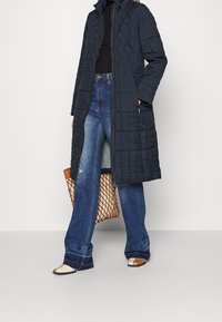 Abrigo largo acolchado azul marino con cuello alto, cierre de cremallera y bolsillos laterales; combinado con jeans acampanados azul claro y zapatos beige.