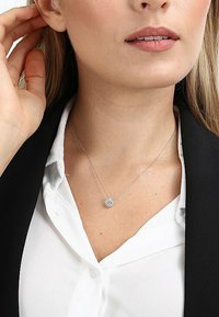 Silbernes Halsband mit einem runden Anhänger, der eine strukturierte, funkelnde Oberfläche aufweist, getragen über einem weißen Hemd unter einem schwarzen Blazer.