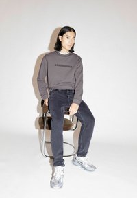 Grijze trui met "ARMEDANGELS" logo, zwarte jeans en zilveren sneakers. Model zit op een chromen stoel met een patroon op de zitting. Neutrale achtergrond.