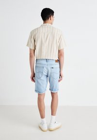 Cămașă cu mâneci scurte, de culoare bej deschis, cu dungi verticale, asortată cu șorturi din denim de culoare albastru deschis. Material texturat, cu margini rulouri și patch cu logo.