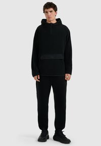 Sweat-shirt noir à capuche avec fermeture éclair partielle, poche avant et poignets élastiques, accompagné d'un pantalon de survêtement noir assorti et de baskets noires.