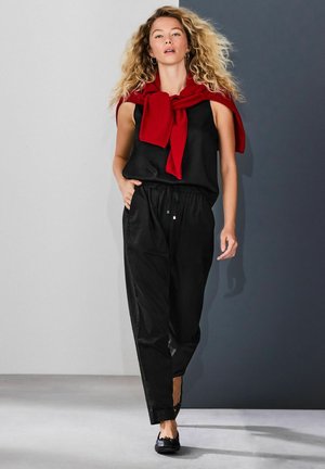Black sleeveless top paired with black trousers featuring ankle slits, accessorized with a red sweater tied at the neck. Black flat shoes.