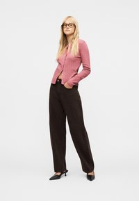 Femme blonde portant des lunettes noires, un cardigan rose boutonné, un pantalon marron à jambes larges et des chaussures noires à petit talon, debout avec une main dans la poche.