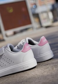 Zapatillas blancas con detalles en rosa, lados perforados, textura suave y una suela texturizada. Presenta el logo de adidas en el talón.