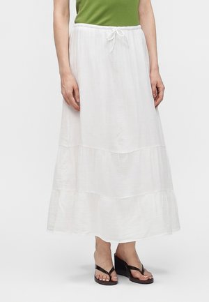 Femme portant une longue jupe blanche à volants avec taille à cordon, haut vert et sandales compensées noires, debout devant un fond uni.
