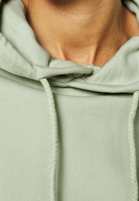 Světle zelená mikina s texturovanou tkaninou, obsahující kapuci se stahovacími šňůrkami a volnější střih. Viditelné švy dodávají detail.