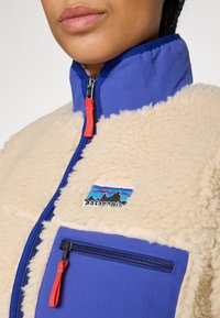 Zbliżenie na polarową kurtkę z niebieskim kołnierzem i kieszenią, beżowy materiał teksturowany, czerwone uchwyty zamka błyskawicznego oraz naszywka z logo Patagonia na klatce piersiowej.
