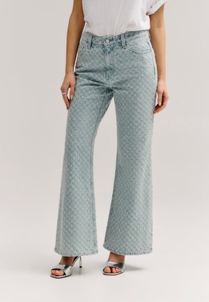 Frau trägt hellblaue, strukturierte, hochtaillierte Schlagjeans mit einem weißen, eingesteckten Hemd und silbernen offenen High-Heels.