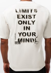 Weißes Baumwoll-T-Shirt mit kurzen Ärmeln. Auf dem Rücken befindet sich auffälliger, schwarz gesprüht Text: "Grenzen existieren nur in deinem Kopf!" sowie eine Weltkugelgrafik.
