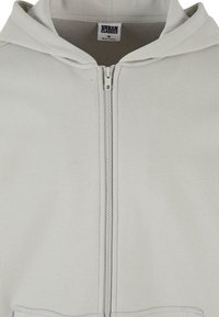 Sweat à capuche gris clair avec fermeture éclair à l'avant, capuche attachée, et étiquette "Urban Classics" à l'intérieur du col, taille moyenne.