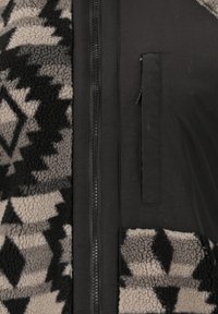 Veste noire avec des panneaux en polaire grise et noire à motifs et une poche zippée verticale à côté de la fermeture éclair centrale à l'avant.