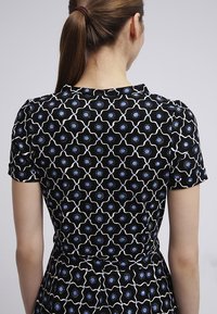 Kobieta z brązowymi włosami spiętymi w kucyk, ubrana w czarną sukienkę z krótkim rękawem ozdobioną niebieskimi i białymi geometrycznymi wzorami kwiatowymi, stojąca tyłem do kamery.
