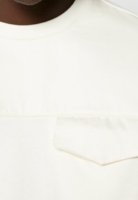 T-shirt bianca in cotone con scollo rotondo e tasca con patta. Il tessuto ha una trama liscia e un leggero effetto lucido.