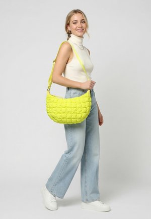 Mujer sonriente con cuello alto sin mangas blanco y vaqueros azul claro sostiene un gran bolso de hombro acolchado amarillo con zapatillas blancas.