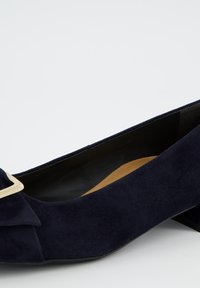 Escarpin en daim bleu marine avec bout rond, petit nœud décoratif et détail en métal doré. Talon bas, solide et noir, avec intérieur lisse.