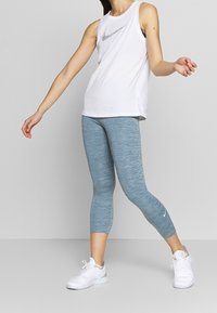 Biała bezrękawnik z srebrnym logo Nike, zestawiony z jasnoniebieskimi melanżowymi legginsami długości kostek i białymi butami sportowymi.