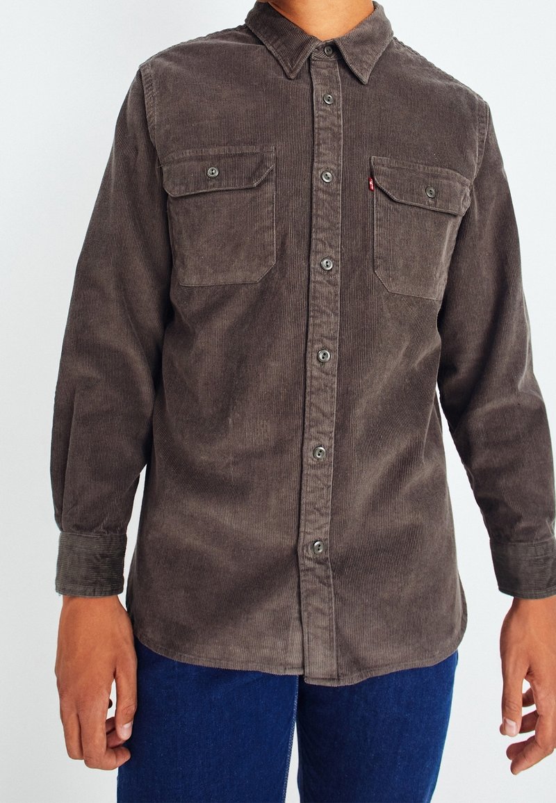 Man in een bruine knoopjescorduroy shirt met twee borstzakken en blauwe broek, staande tegen een effen achtergrond.