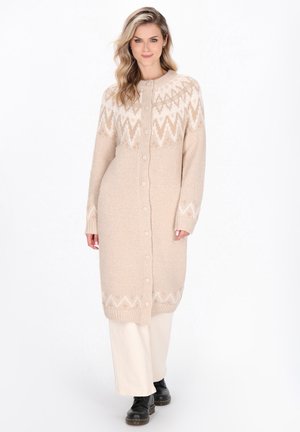 Beige gebreide cardigan met een zigzagpatroon in lichtere tinten. Heeft knopen aan de voorkant en lange mouwen, gedragen over crèmekleurige broeken.