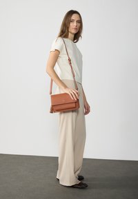 Tan leather crossbody bag with a flap and tassel detail. Worn on a model paired with a cream top and beige wide-leg trousers.