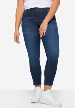 Jean en denim bleu foncé avec une coupe slim, présentant des poches avant, une fermeture à bouton et à zip, ainsi que des détails de couture subtils. Associé à des baskets montantes blanches.