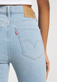 Ljusblå denimjeans med hög midja, med en texturerad brun etikett på baksidan och en röd Levi's etikett på fickan.