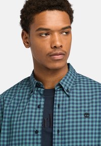 Camicia a quadri verde e blu con colletto button-down, due bottoni neri e un piccolo logo ricamato sul petto. Tessuto dalla texture liscia.