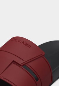 Μαύρο σανδάλι με φαρδιά σκουρόκοκκινη λουρίδα που φέρει ανάγλυφο λογότυπο Calvin Klein και λεπτομέρεια με κοψίματα.