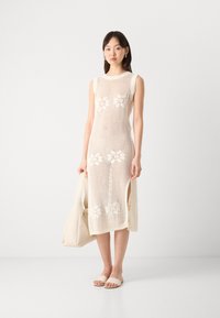 Knit dress in cream with floral cutouts, sleeveless design, side slits, paired with a light bag and neutral sandals, on a plain background.