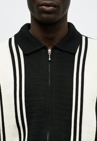 Κοντινό πλάνο ενός ατόμου που φοράει μαύρο και άσπρο πλεκτό πουκάμισο με φερμουάρ, κάθετες ρίγες και γιακά, εμφανίζοντας μόνο τον λαιμό και το κάτω μέρος του προσώπου.