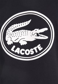 Białe logo Lacoste z otwartym krokodylem wewnątrz dwóch koncentrycznych okręgów na czarnym tle.
