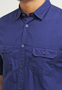 Mörkblå kortärmad knäppskjorta i bomull, med två bröstfickor, krage och blå knappar. Slen textur med en avslappnad passform.