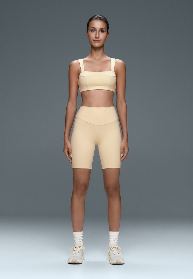 Femme debout face à l’avant, portant un soutien-gorge de sport beige, un short de cyclisme taille haute, des chaussettes blanches et des baskets beiges sur un fond gris uni.
