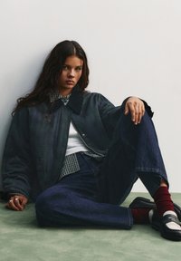 Dunkelgrüne gesteppte Jacke mit Cordkragen, über einem karrierten Hemd getragen. Dunkelblaue weit geschnittene Jeans und schwarze Loafer mit roten Socken.