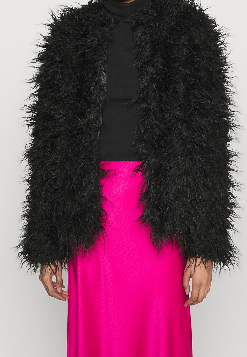 Personne portant une veste noire texturée et duveteuse sur un haut noir et une jupe en satin rose vif, debout devant un fond uni.