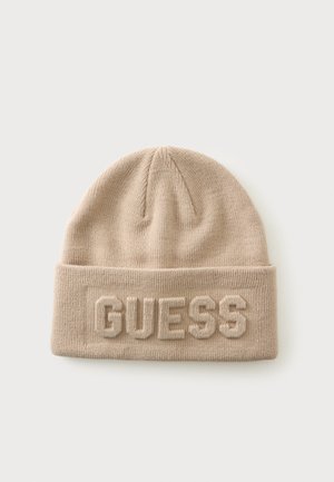 Beżowa dziana czapka z podwiniętym mankietem z wypukłymi literami "GUESS" z przodu, na jednolitym jasnym tle.