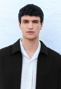 Melna teksturēta jaka ar rāvējslēdzēju, valkāta virs baltas krekla ar sarežģītu mežģīņu izšuvumu un pogām, attēlota uz gaismas fona.