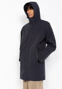 Chaqueta impermeable azul marino con capucha, con un corte recto, dos bolsillos laterales y cordones ajustables. Textura suave con un diseño minimalista.