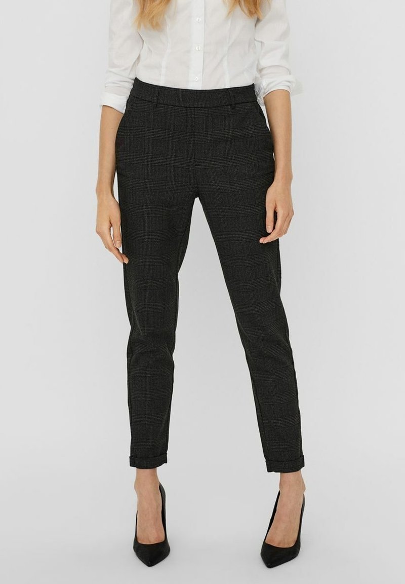 Femme portant un pantalon slim à carreaux gris foncé avec des ourlets retroussés, associé à des escarpins noirs à bout pointu et une chemise blanche boutonnée.