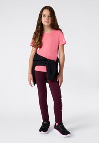 Fiatal lány, hosszú, göndör hajjal, rózsaszín pólót, bordó leggingset, fekete sportcipőt visel, és egy fekete pulóvert kötött a derekára.