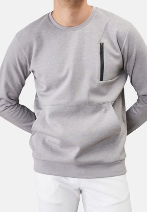 Uomo che indossa una felpa a maniche lunghe grigio chiaro con una tasca verticale con cerniera nera sul petto e pantaloni bianchi su uno sfondo uniforme.