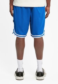 Pantalones cortos deportivos azules con ribetes blancos y aberturas laterales, hechos de material transpirable, usados con calcetines blancos y zapatos negros, vistos desde detrás.