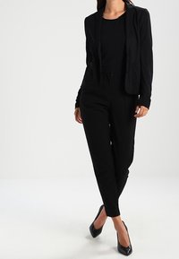 Blazer nero e pantaloni abbinati realizzati in tessuto morbido. Il blazer presenta due tasche frontali e una chiusura con un solo bottone.