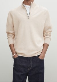 Man in beige quarter-zip sweater over white shirt with hands in dark blue jeans pockets, wearing a silver bracelet on left wrist.