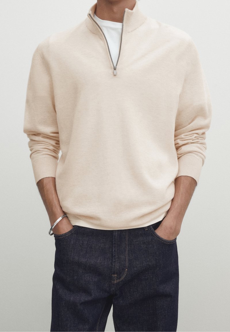 Man in beige quarter-zip sweater over white shirt with hands in dark blue jeans pockets, wearing a silver bracelet on left wrist.
