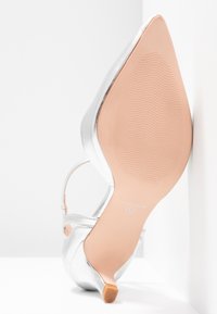 Chaussure à talon aigu en argent avec un bout pointu, finition métallique, fine sangle à la cheville et semelle beige texturée. Design élégant et raffiné.