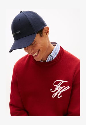 Jeune homme souriant, portant une casquette de baseball marine et un pull rouge avec les initiales blanches "TH" sur une chemise à col rayée bleue.