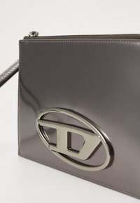 Clutch in pelle sintetica grigia con un prominente logo ovale argento. Presenta una chiusura con zip e una consistenza morbida, arricchita da dettagli di cucitura.