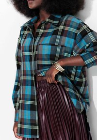 Chemise à carreaux bleue et noire avec des manches longues, col boutonné et une poche, assortie à une jupe plissée de couleur bourgogne foncé.